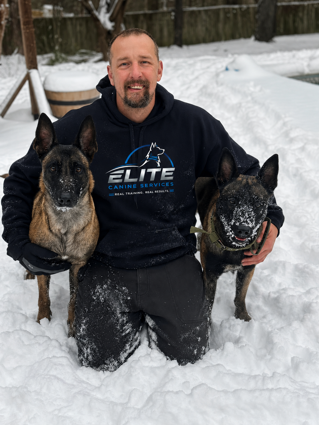 Alan kneeling in the snow with two Belgian Malinois while wearing a Sit Means Sit hoodie
